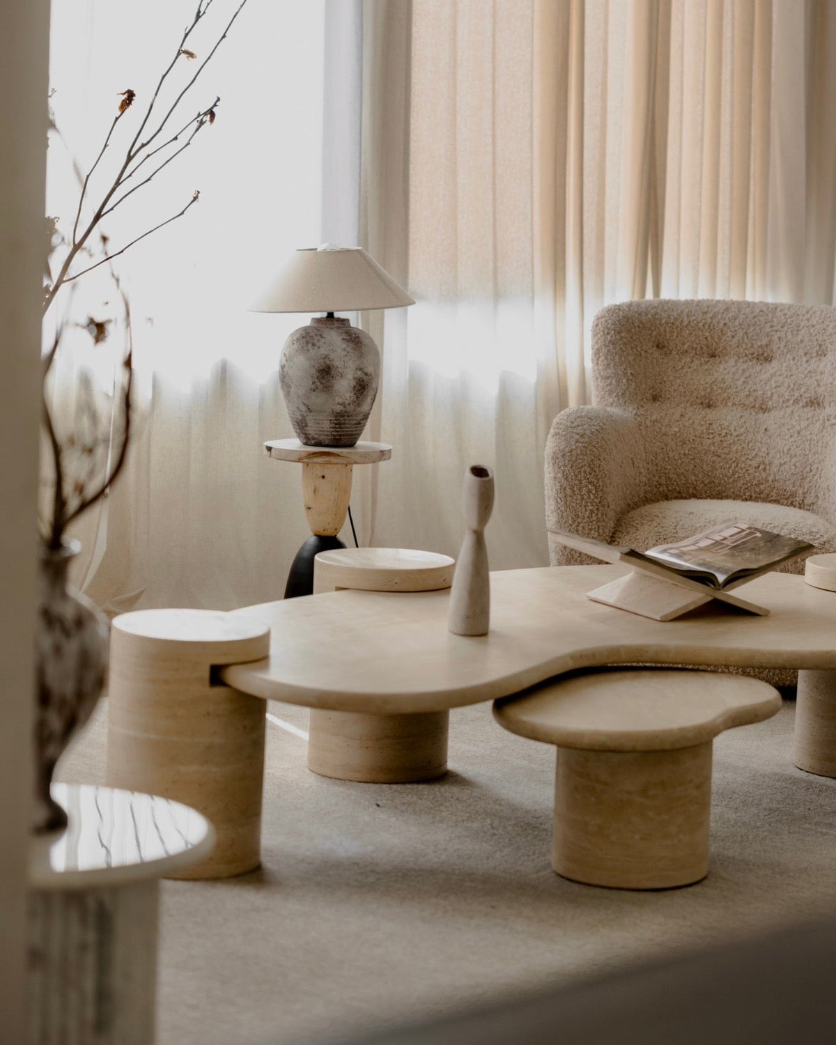 Tuscani travertine kidney coffee table with a nesting side table, beside a boucle armchair and a ceramic lamp in soft window light